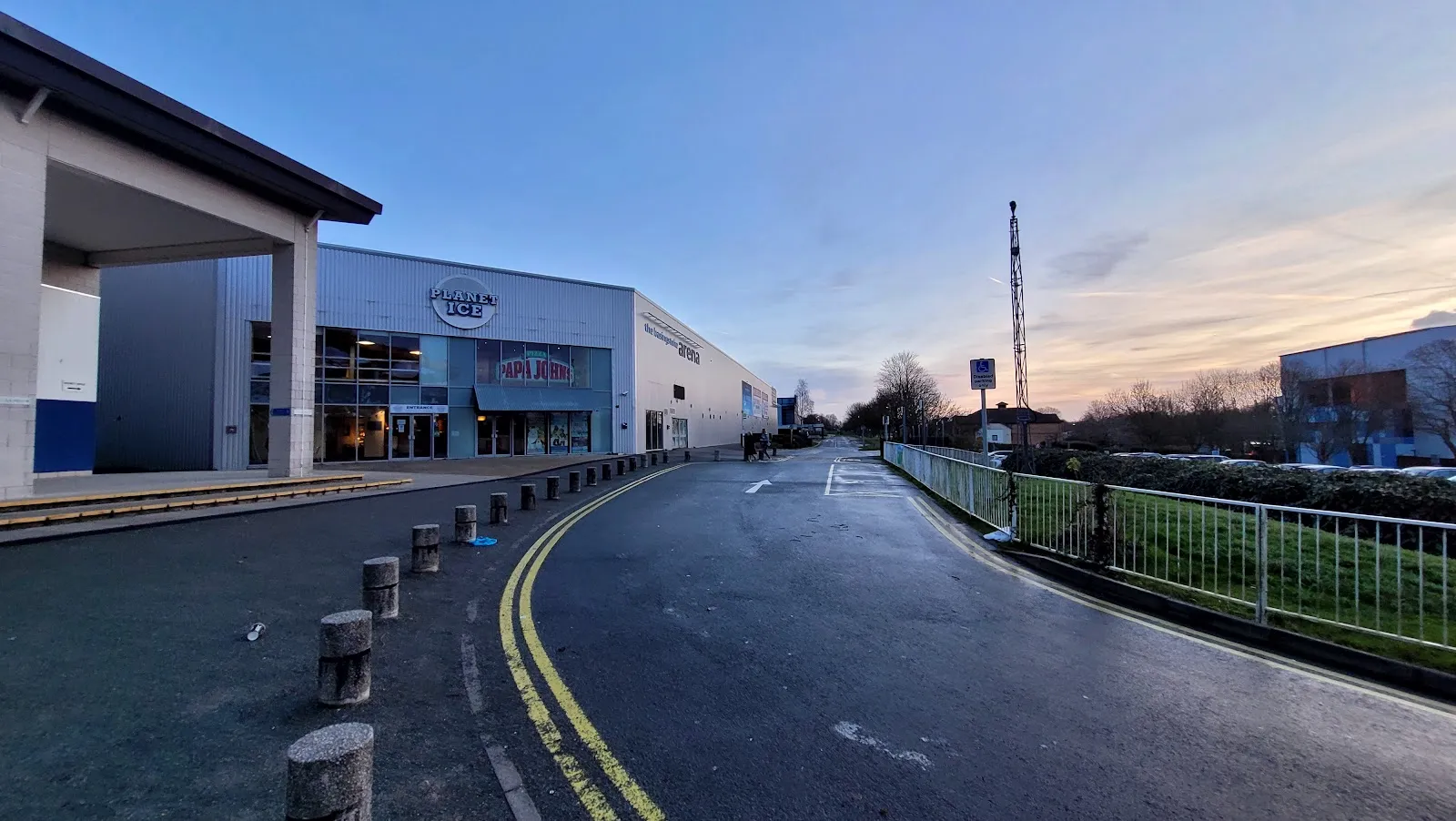 The grey cladded Basingstoke arena at dusk from the access road, with the Planet Ice sign above the glazed entrance and 'the basingstoke arena' lettering along the side. — Planet Ice Basingstoke