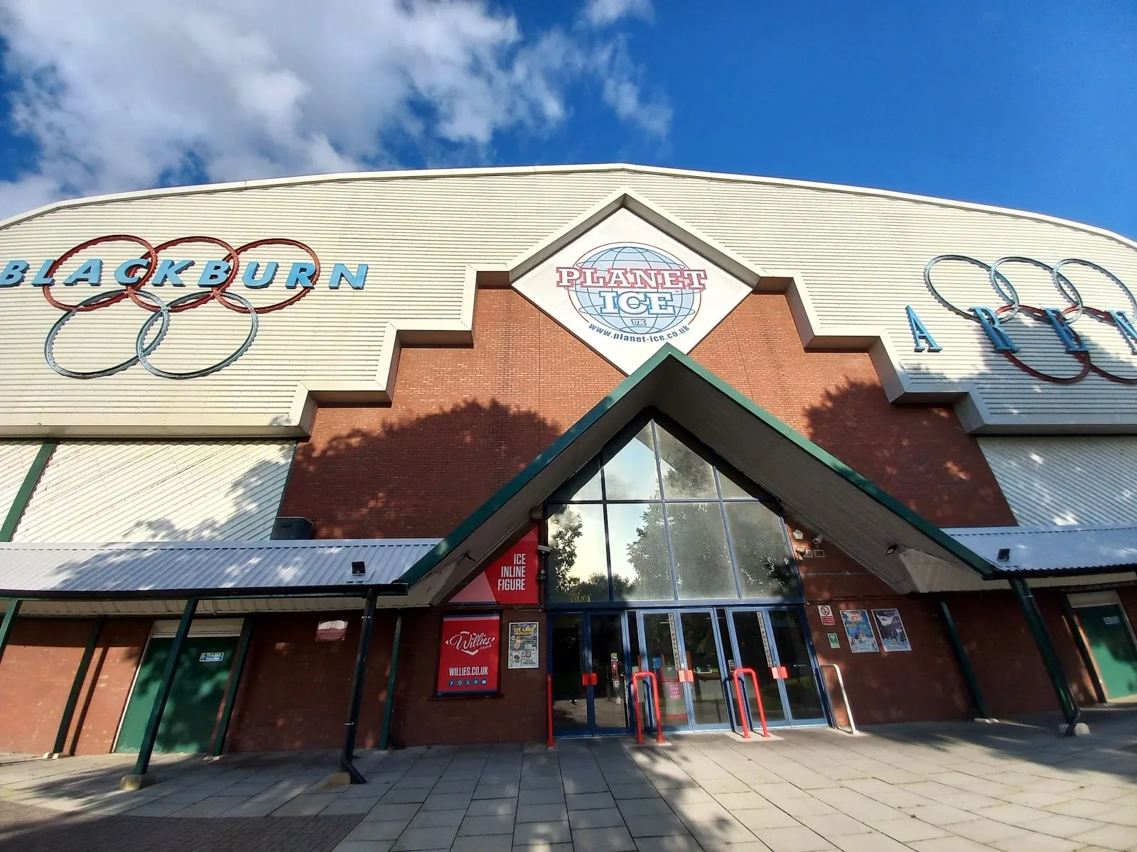 The front of Blackburn Arena: the brick entrance gable with the Planet Ice globe, the glass doors below and the 'Blackburn Arena' lettering and Olympic rings on the cladding. — Planet Ice Blackburn