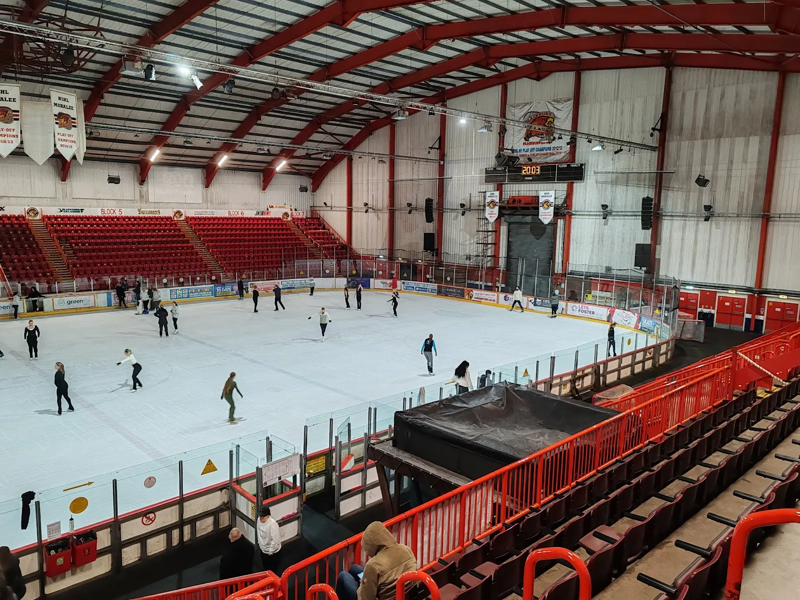 The ice sheet from a high seat during a public session, skaters spread across it, red steel arches over the stands on the far side. — Planet Ice Blackburn