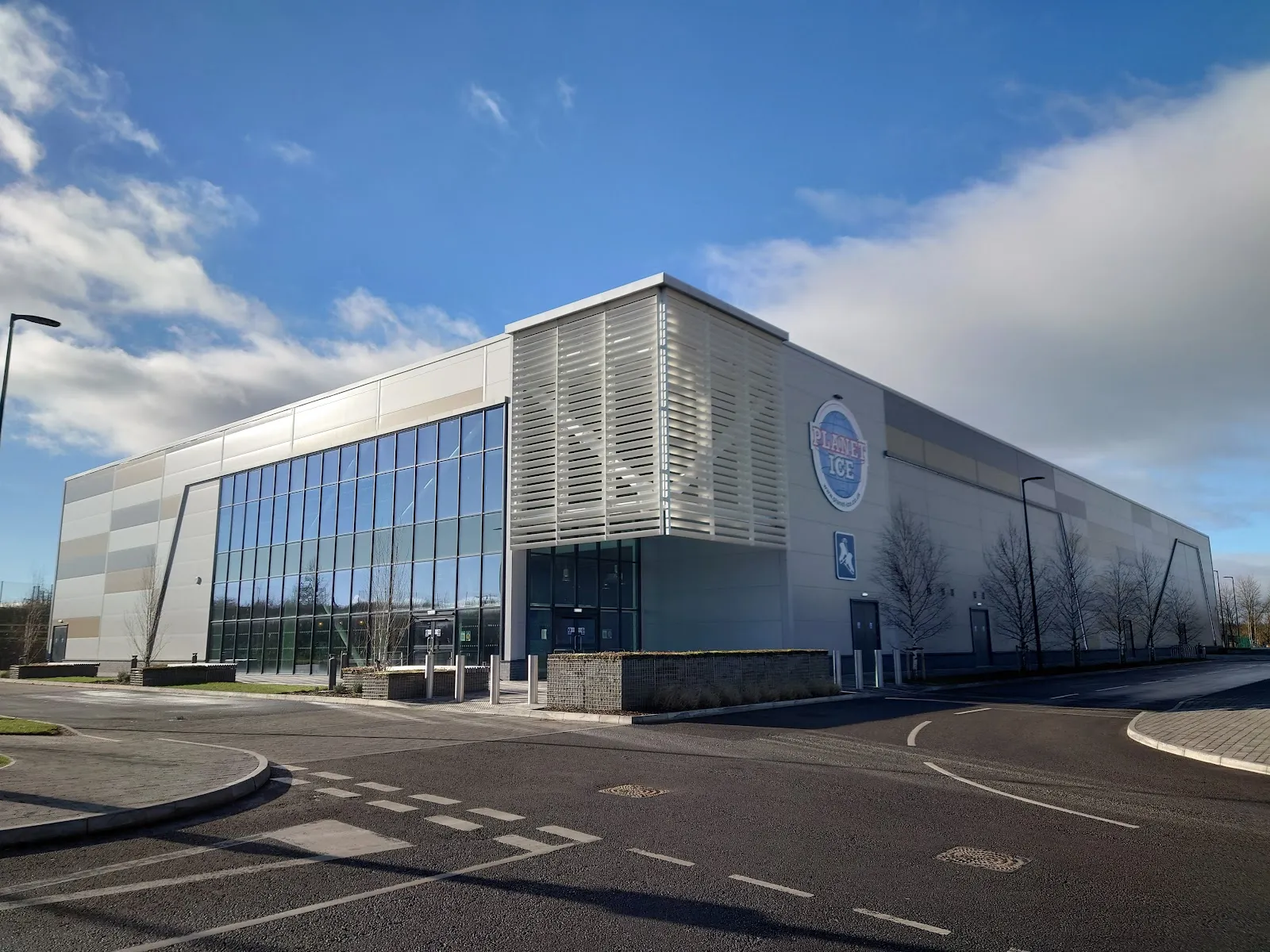 The new glass-fronted arena with grey cladding and the Planet Ice globe on the end wall, seen from the access road under a blue sky. — Planet Ice Bristol
