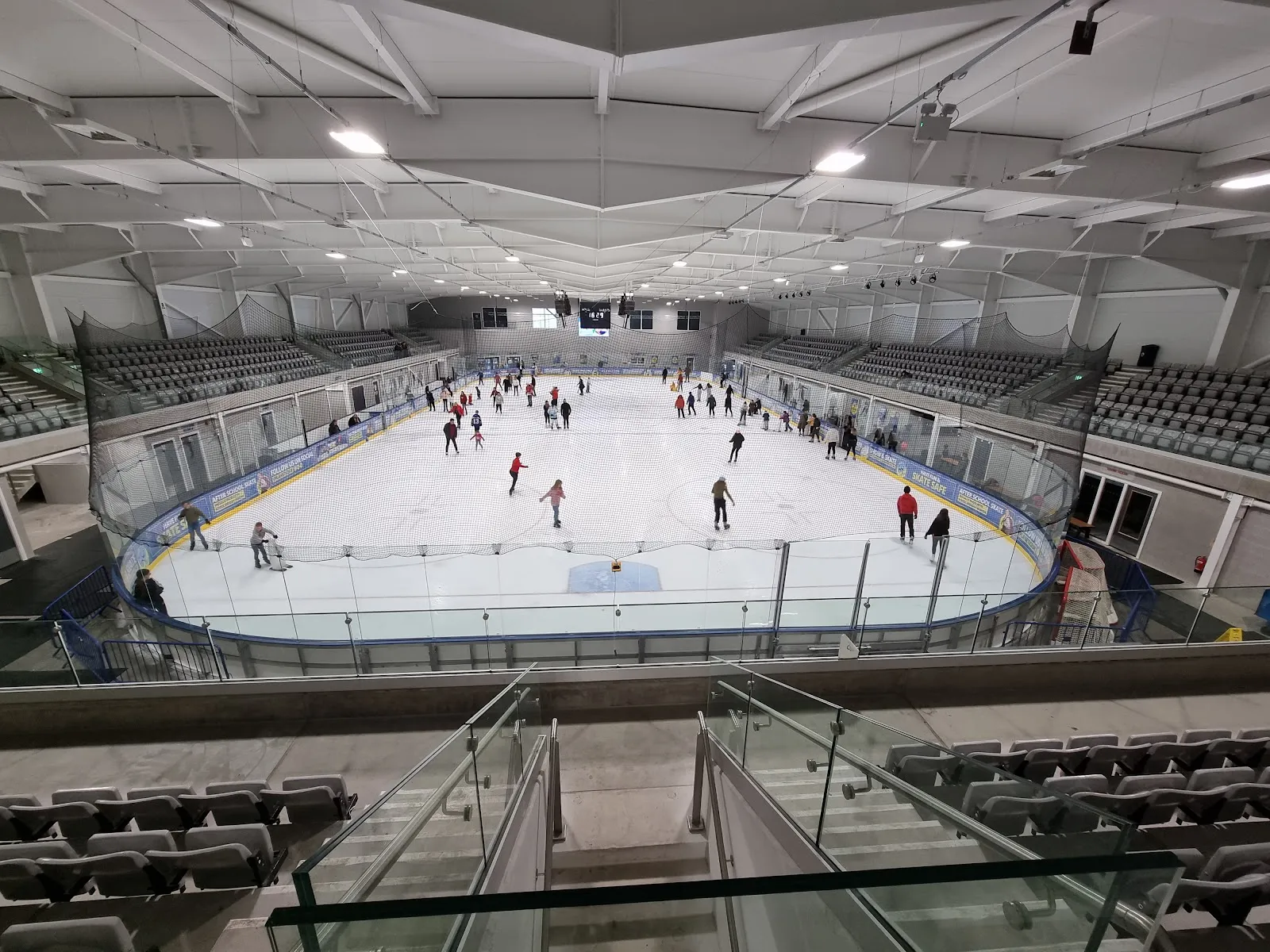 The whole ice sheet seen from the upper concourse, with skaters on it at a public session and seating banks and a steel roof around. — Planet Ice Bristol