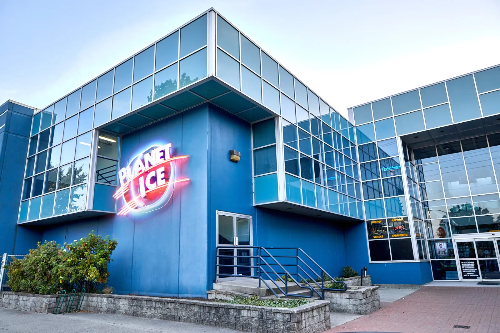 The blue glass-and-steel entrance with the neon Planet Ice sign, landscaped planters and the brick walkway to the front doors. — Planet Ice Coquitlam