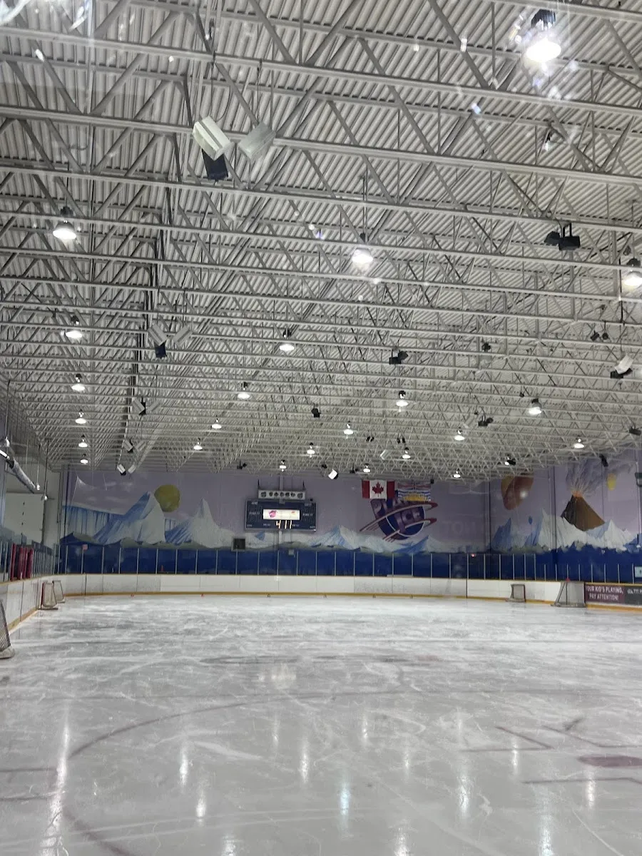 An empty rink under white steel trusses, with a mountain mural and the Planet Ice logo painted along the far wall. — Planet Ice Coquitlam