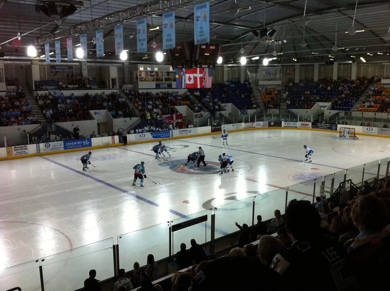 An ice hockey game under way in the Skydome Arena, the Coventry Blaze at a face-off with banners hung from the roof trusses and fans in the stands. — Planet Ice Coventry
