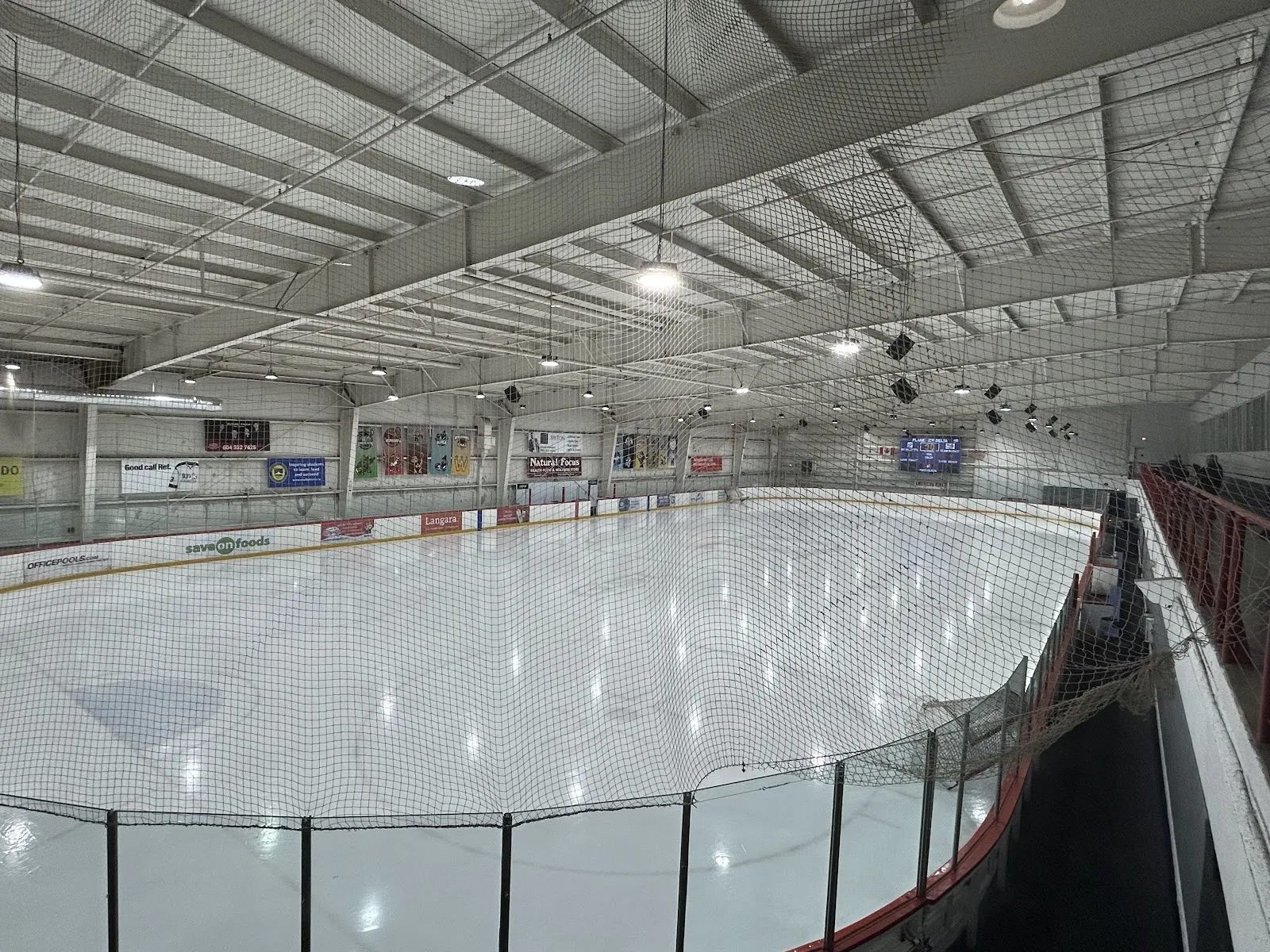 An empty rink seen from the stands behind netting, with sponsor boards along the far boards and a low steel roof. — Planet Ice Delta