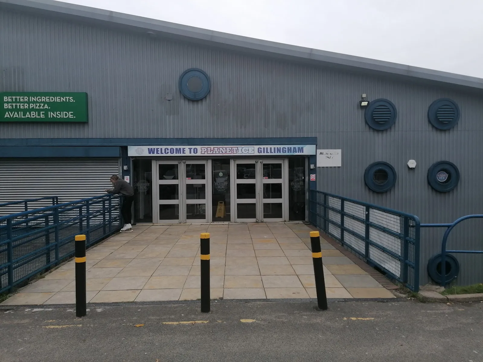 The grey clad hall seen from the car park side, with the 'Welcome to Planet Ice Gillingham' sign above the double glass entrance doors. — Planet Ice Gillingham