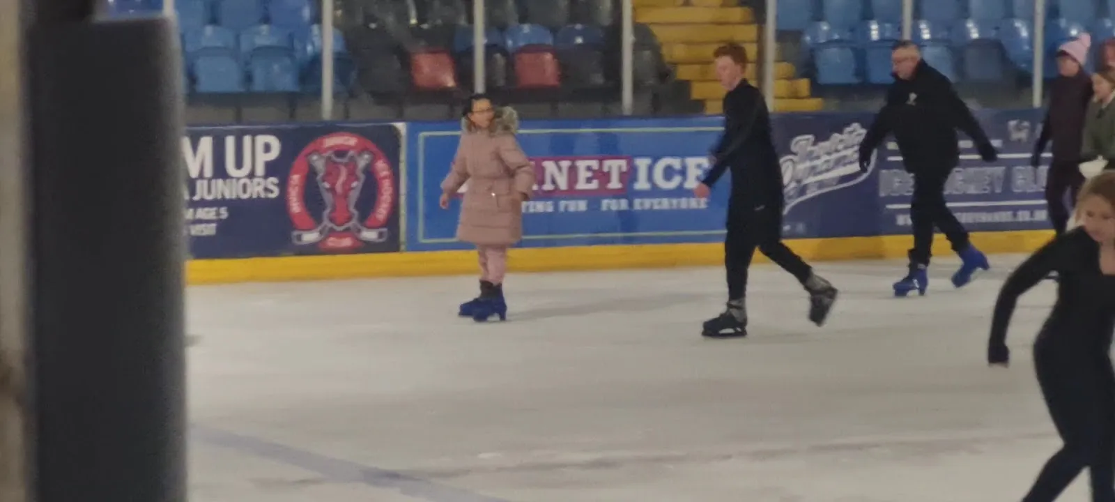 Skaters crossing the ice along the sponsor boards at Planet Ice Gillingham, with the blue and red seating behind the glass. — Planet Ice Gillingham