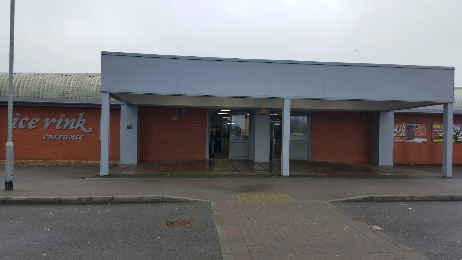 The low brick-and-grey entrance block of the Gosport rink on a grey day, with the 'ice rink entrance' lettering on the brick wall to the left and glass doors under a flat canopy. — Planet Ice Gosport