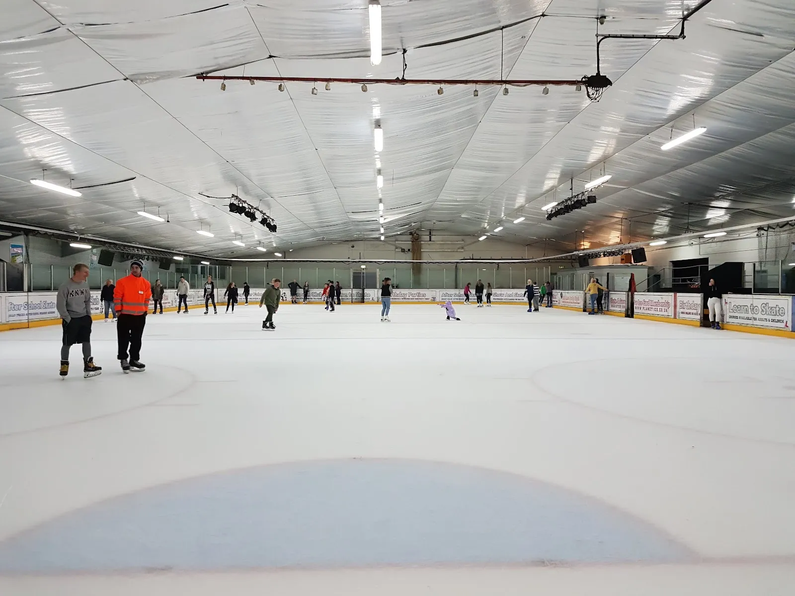 The full ice sheet during a public session, a handful of skaters spread across it, advertising boards around the sides and a white curved ceiling. — Planet Ice Gosport