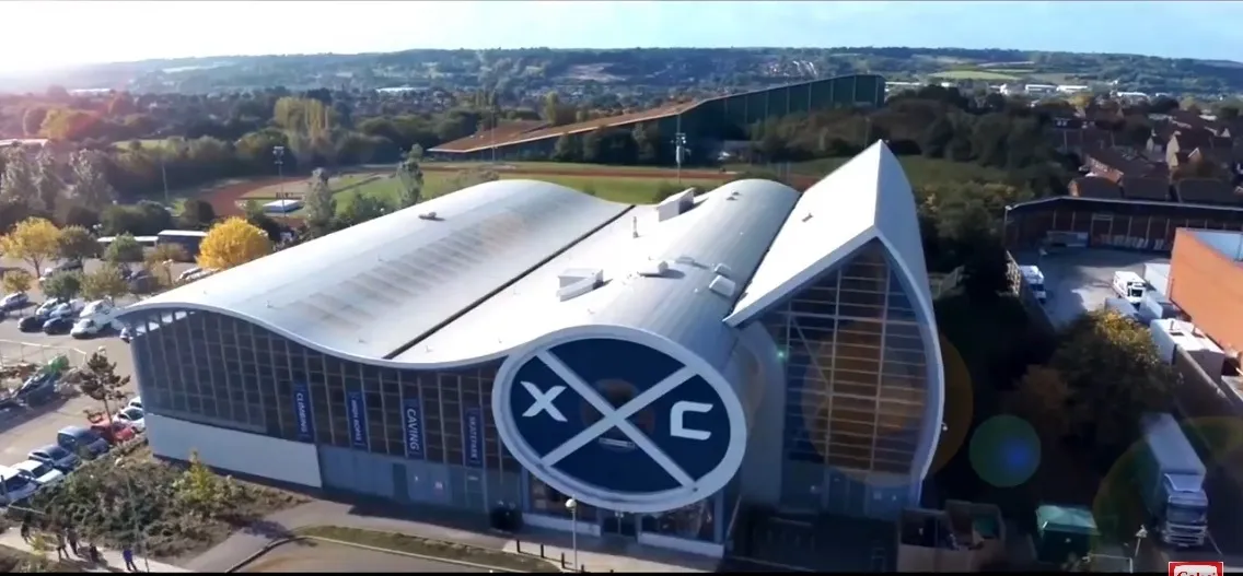 An aerial view of the Hemel Hempstead Planet Ice building with its curved white and blue roof and crossed-hockey-stick logo on the facade. — Planet Ice Hemel Hempstead