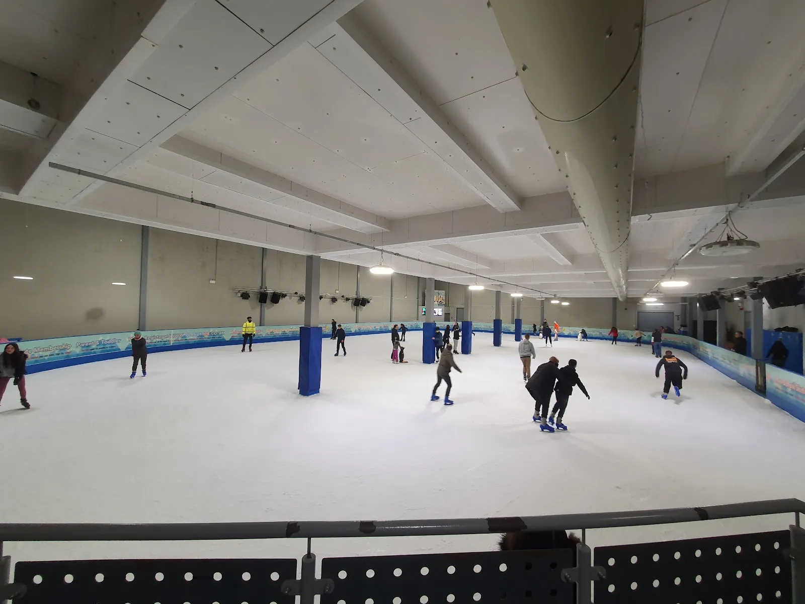 The ice sheet from the barrier during a public session, a scatter of skaters under a low white ceiling with blue pillar pads. — Planet Ice Hemel Hempstead