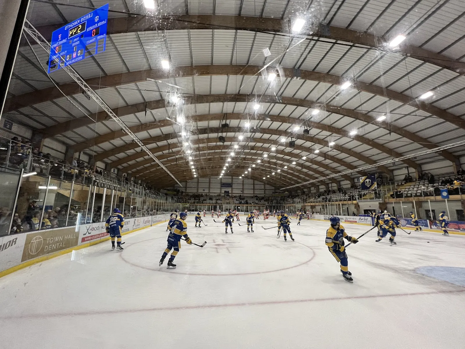 The wide Planet Ice Leeds rink under its curved timber roof and white lighting, skaters spread across the ice with the boards and seating beyond. — Planet Ice Leeds