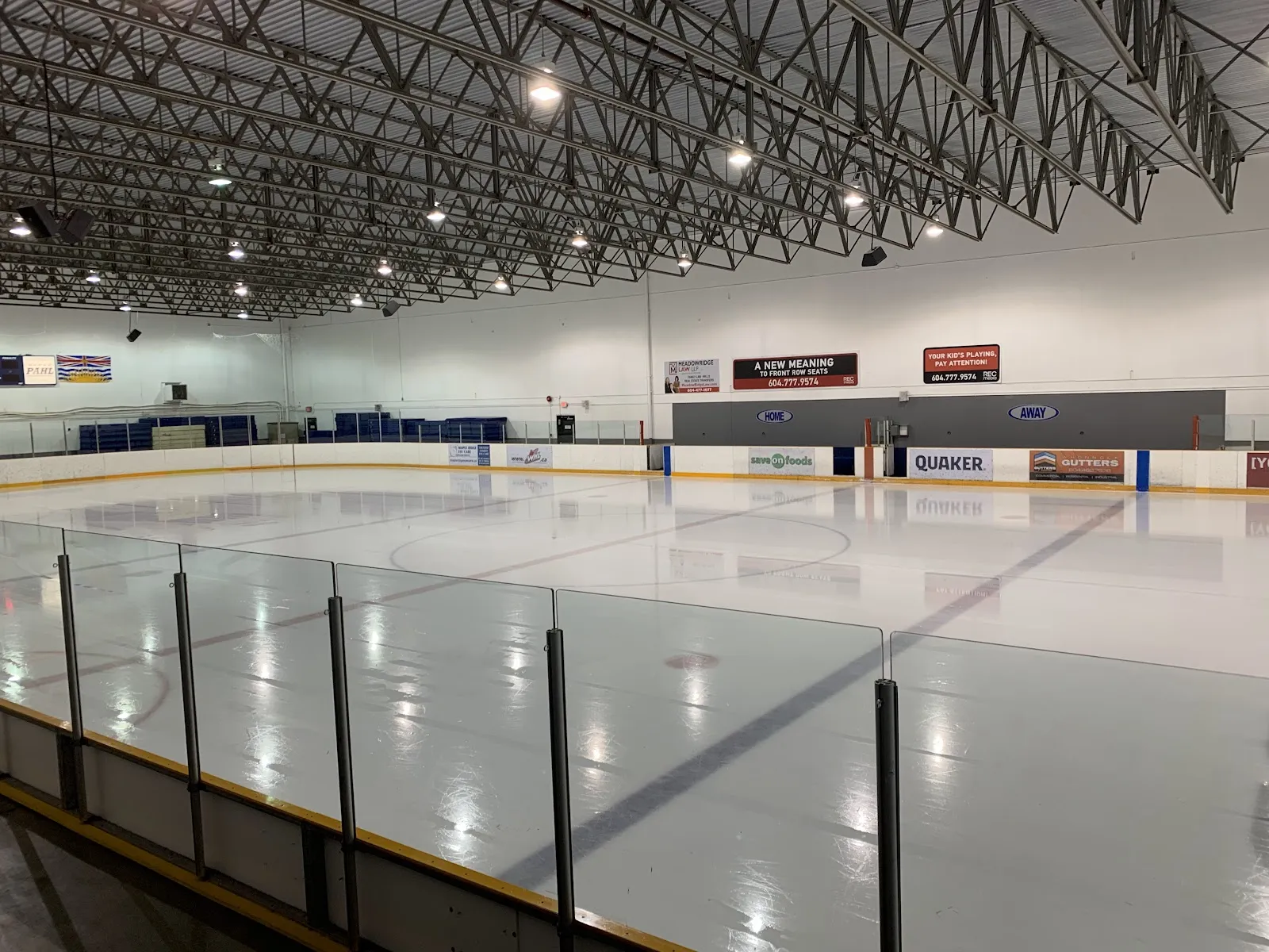 An empty rink seen over the glass from the end stands, with dark steel trusses and sponsor boards on the far side. — Planet Ice Maple Ridge