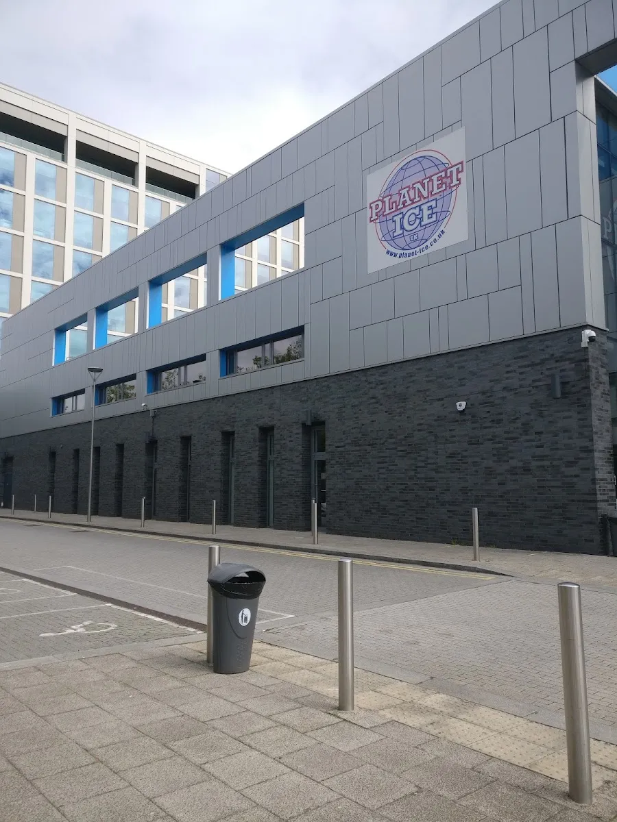 The side wall of the grey cladded arena with the Planet Ice globe sign and a row of blue-framed windows, an office block behind. — Planet Ice Milton Keynes