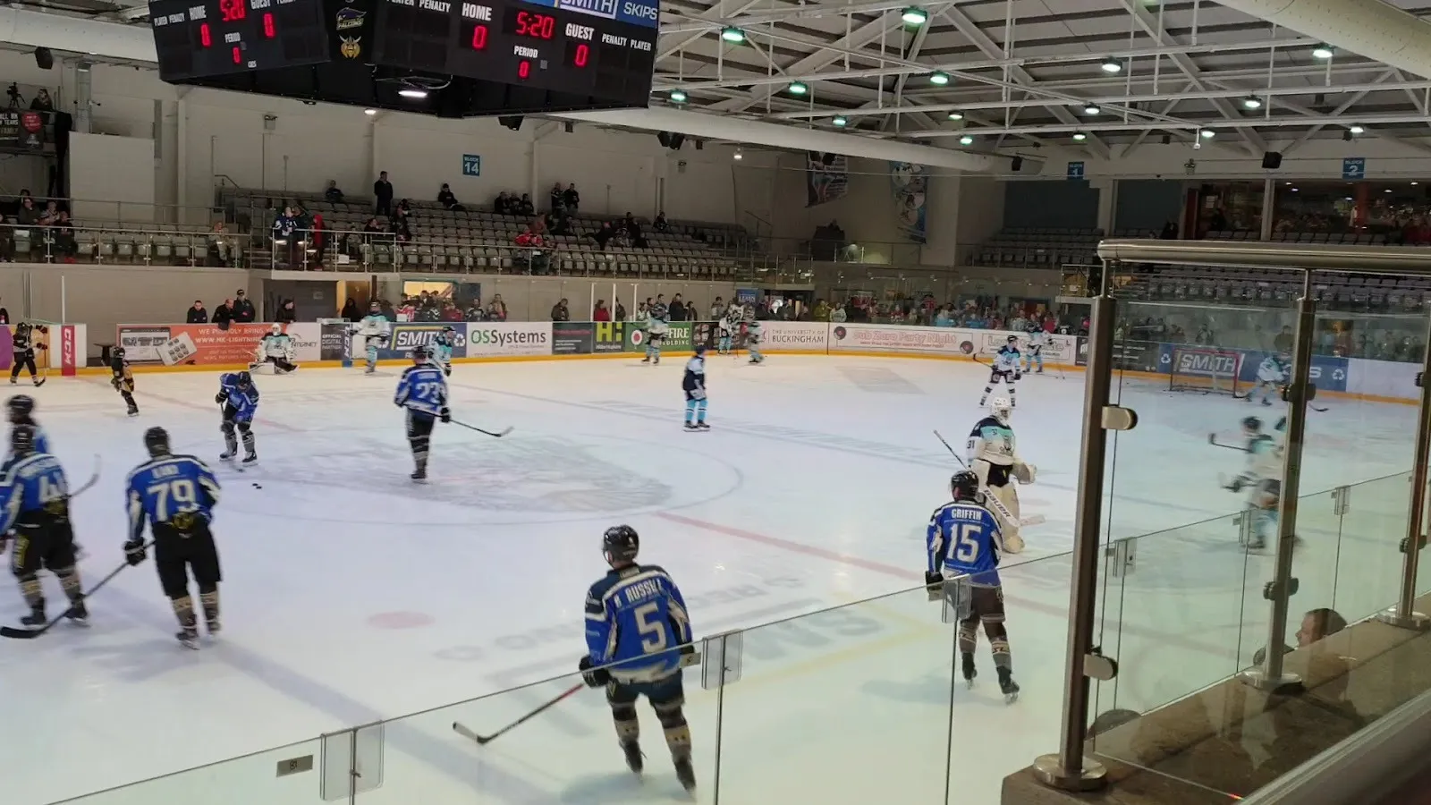 An ice hockey game in progress at Planet Ice Milton Keynes, players in blue chasing the puck across the rink with spectators behind the glass and a scoreboard overhead. — Planet Ice Milton Keynes