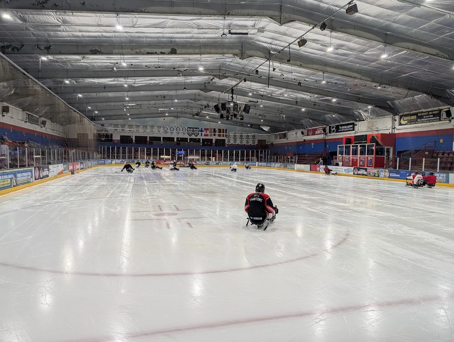 The wide Planet Ice Peterborough rink under its low steel roof, the centre line and face-off circle marked on the ice with a sledge hockey player and a few skaters at the far end. — Planet Ice Peterborough