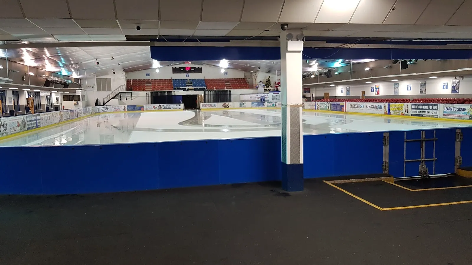 The empty ice sheet seen across the rink barrier, with the blue boards, the far stands and the roof lights. — Planet Ice Solihull