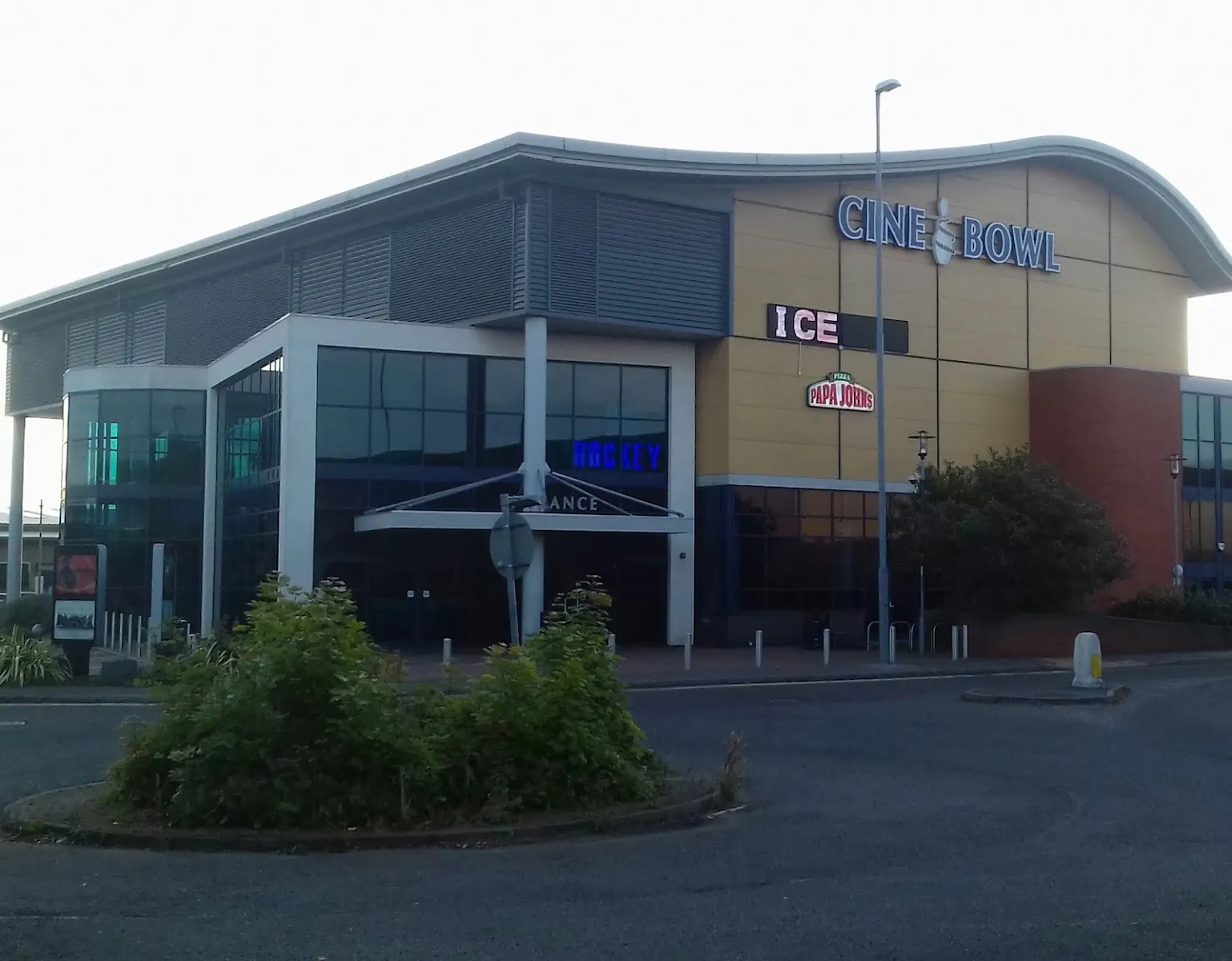 The curved-roof CineBowl building on Meadows Way, with the lit 'CineBowl' sign and a separate 'ICE' sign above the glazed entrance, seen from the access road at dusk. — Planet Ice Uttoxeter
