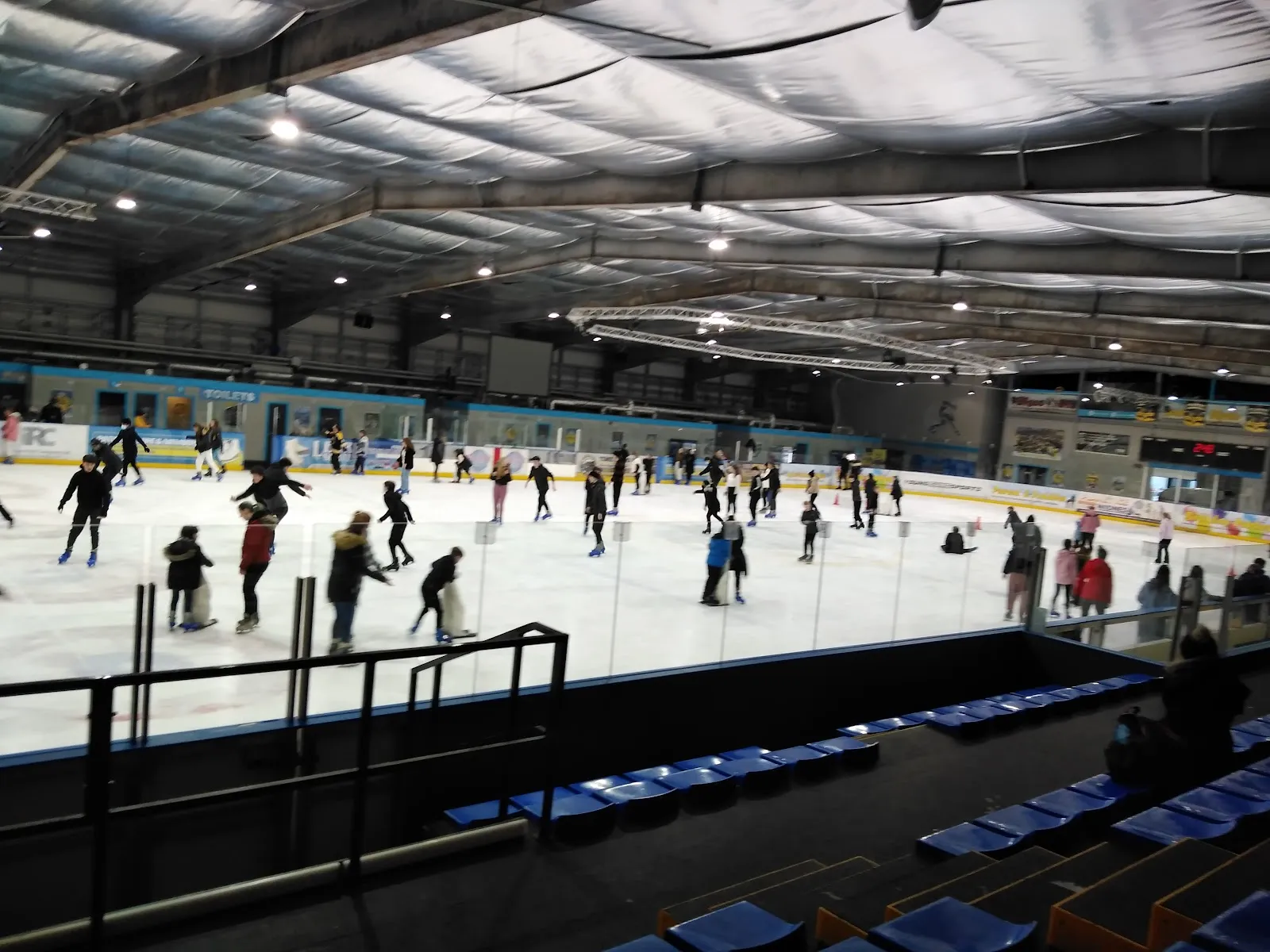 The ice sheet from the raised seating during a public session, skaters spread across it and the far boards and roof beyond. — Planet Ice Widnes