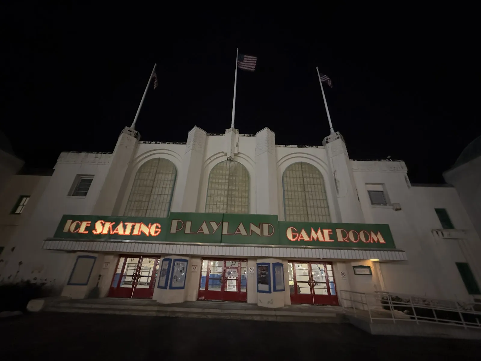Vista nocturna de la fachada frontal del edificio Art Déco de Playland, iluminada con reflectores, con tres astas de bandera arriba y una marquesina que dice 'ICE SKATING PLAYLAND GAME ROOM' sobre las puertas de la entrada. — Playland Ice