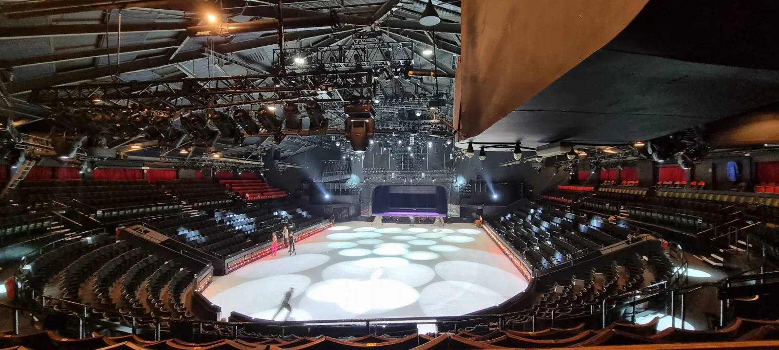 The ice in the Pleasure Beach Arena theatre, lit by round spotlights, with a few skaters and tiered seating on all sides (cropped). — Pleasure Beach Arena