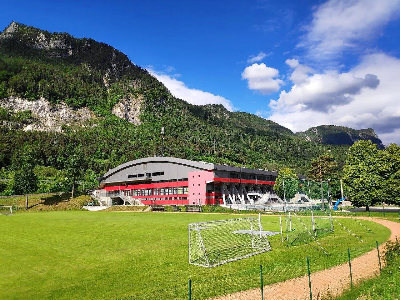 The Podmežakla Sports Hall in Jesenice, a curved-roof hall with a red and grey facade, seen across a football pitch with the Karavanke slopes rising behind. — Podmežakla Sports Hall