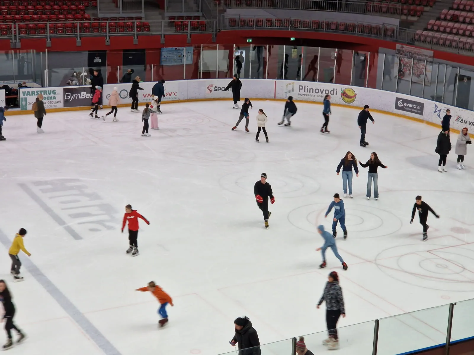 Skaters of all ages circling the ice of the Podmežakla Sports Hall during a recreational skating session, seen from the stands. — Podmežakla Sports Hall