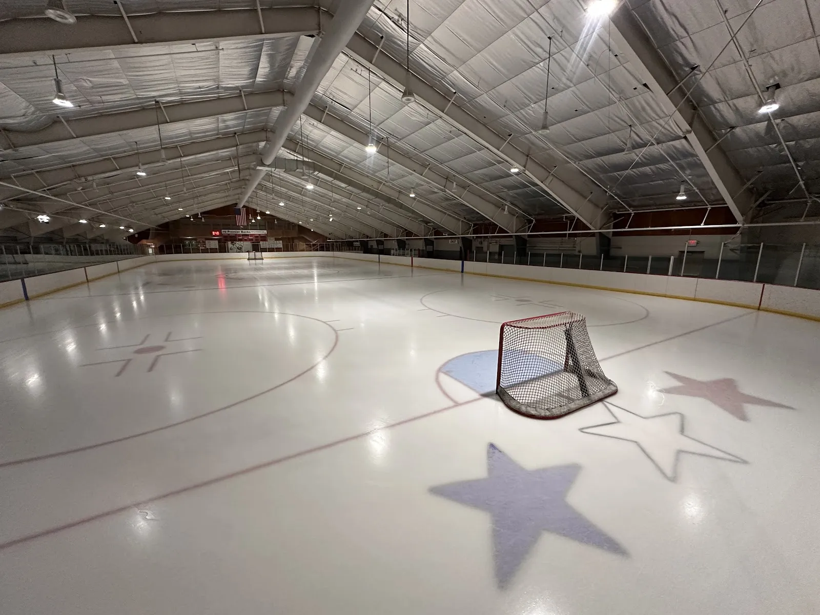 Pista de hielo vacía bajo el techo en arco forrado de aislante, con una portería de hockey vacía en el centro de la pista, que tiene grandes estrellas pintadas, y vallas y gradas a los lados, sin personas visibles. — Polar Arena (ISD 622)
