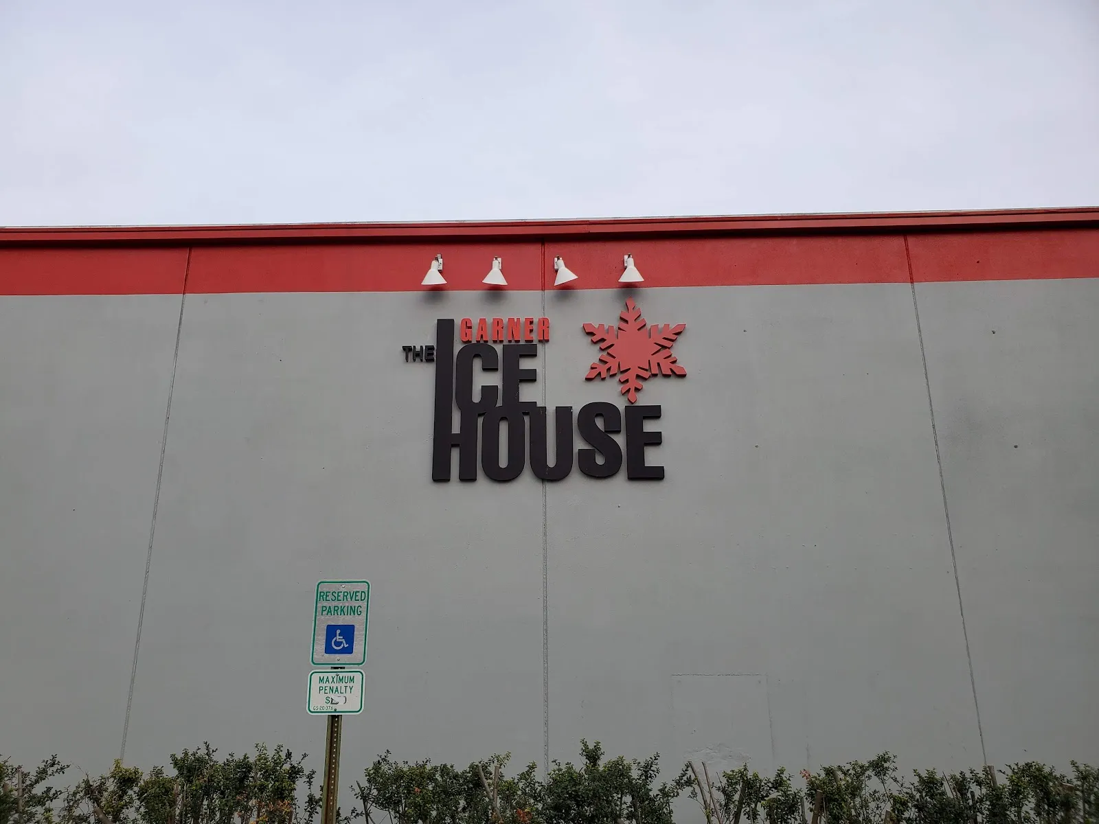 The grey outside wall with red trim and The Garner Ice House sign, above a hedge and an accessible parking sign. — Polar Ice Garner