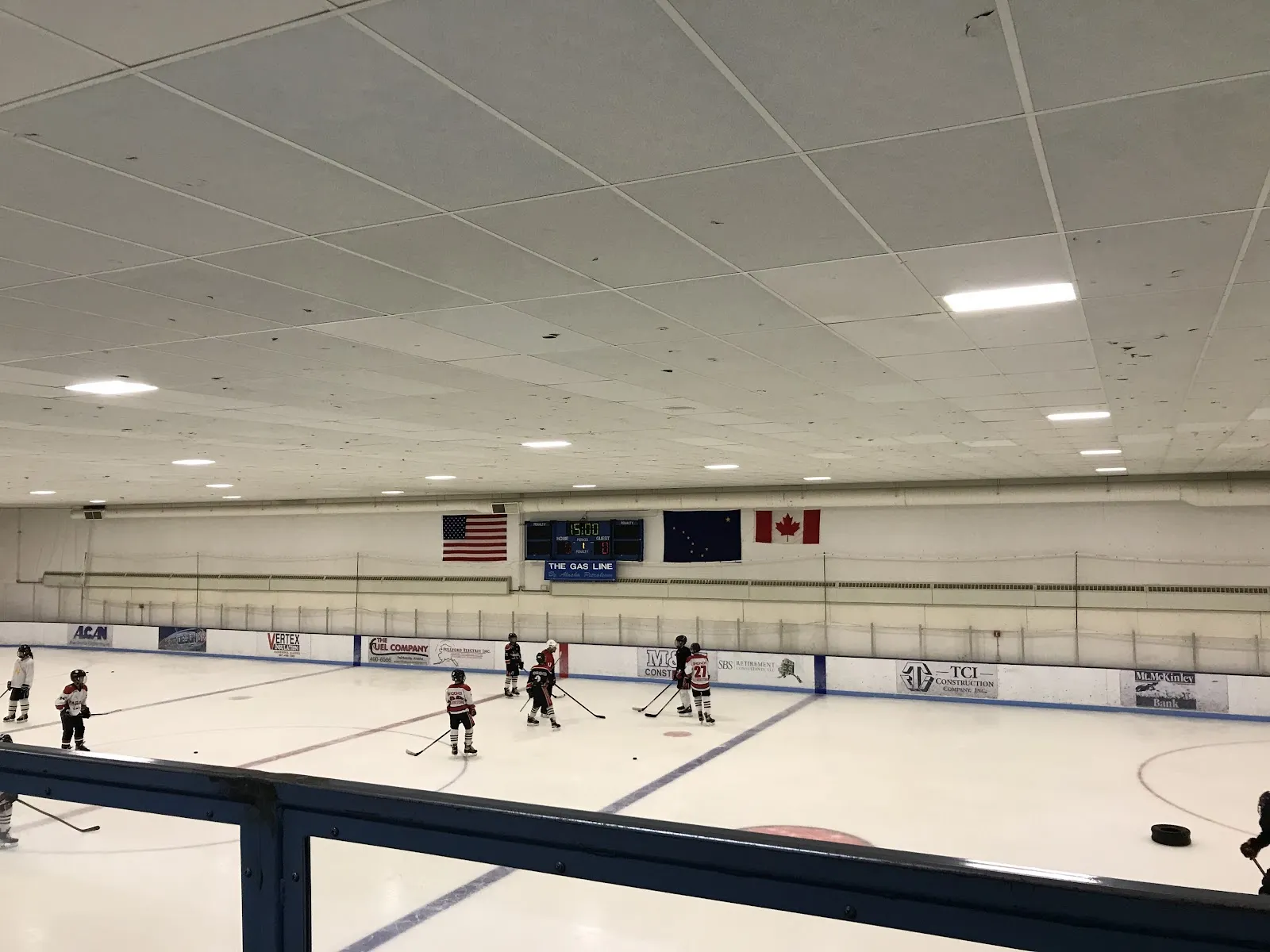 Amplia vista de la pista desde las gradas, con banderas de EE. UU., del estado de Alaska y de Canadá y anuncios de patrocinadores locales a lo largo de las vallas, y jugadores juveniles de hockey patinando a lo lejos. — Polar Ice Center