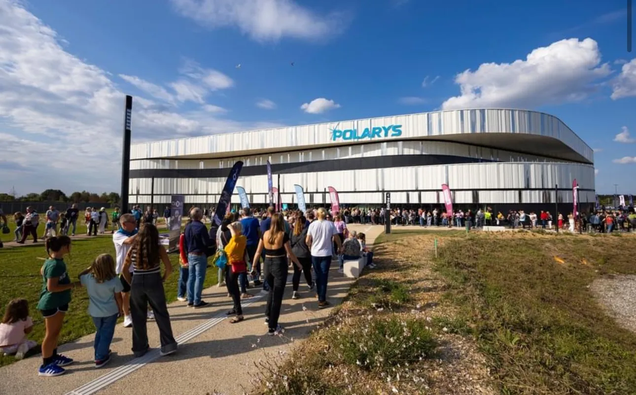 Le long bâtiment blanc de la patinoire Polarys avec son toit courbe et son nom sur la façade, des visiteurs rassemblés sur l’herbe devant. — Patinoire Polarys