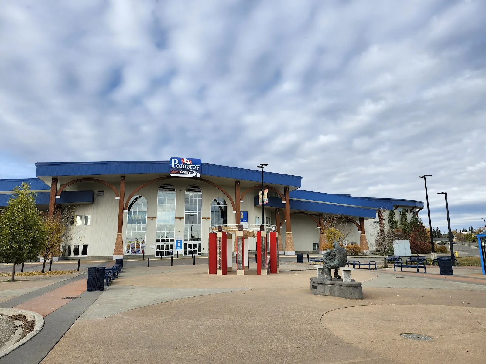 The front of the Pomeroy Sport Centre across its paved plaza, with the blue canopy, the Pomeroy sign above the arched windows and a bronze statue beside the entrance. — Pomeroy Sport Centre