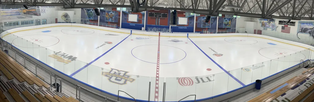 Toda la pista de tamaño NHL vista desde las gradas, con vallas rojas y azules con las bancas HOME y VISITOR, logotipos de equipos pintados en el hielo y mantas de patrocinadores en el muro del fondo, y una máquina pulidora de hielo estacionada en el centro de la pista. — The Ponds of Brookfield