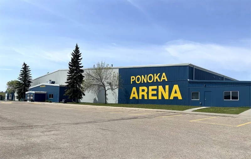 The blue Ponoka Arena building with PONOKA ARENA painted in yellow on its side, a parking lot and spruce trees in front. — Ponoka Arena Complex