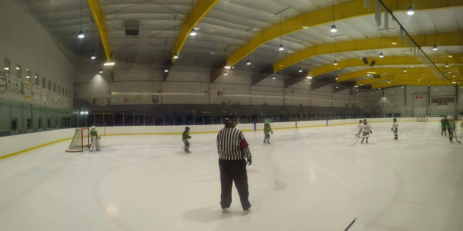 A game seen across a wide sheet with a referee in the foreground, players far away under a blue-trimmed roof with yellow steel braces. — Ponoka Arena Complex