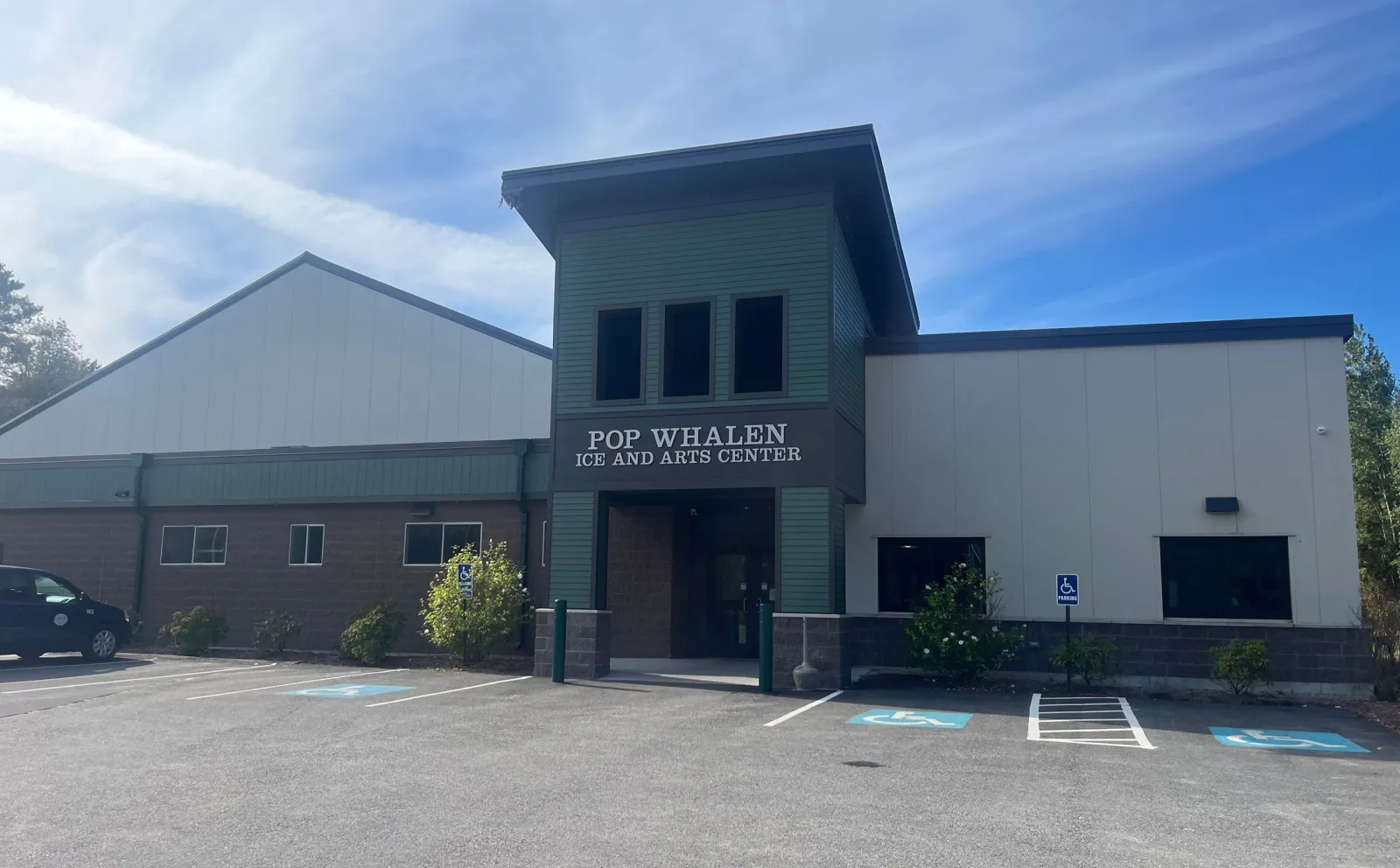 Front entrance of the Pop Whalen Ice and Arts Center, a green-and-tan metal building with a gabled arena roof behind the entry tower, seen from the parking lot on a sunny day. — Pop Whalen Ice & Arts Center