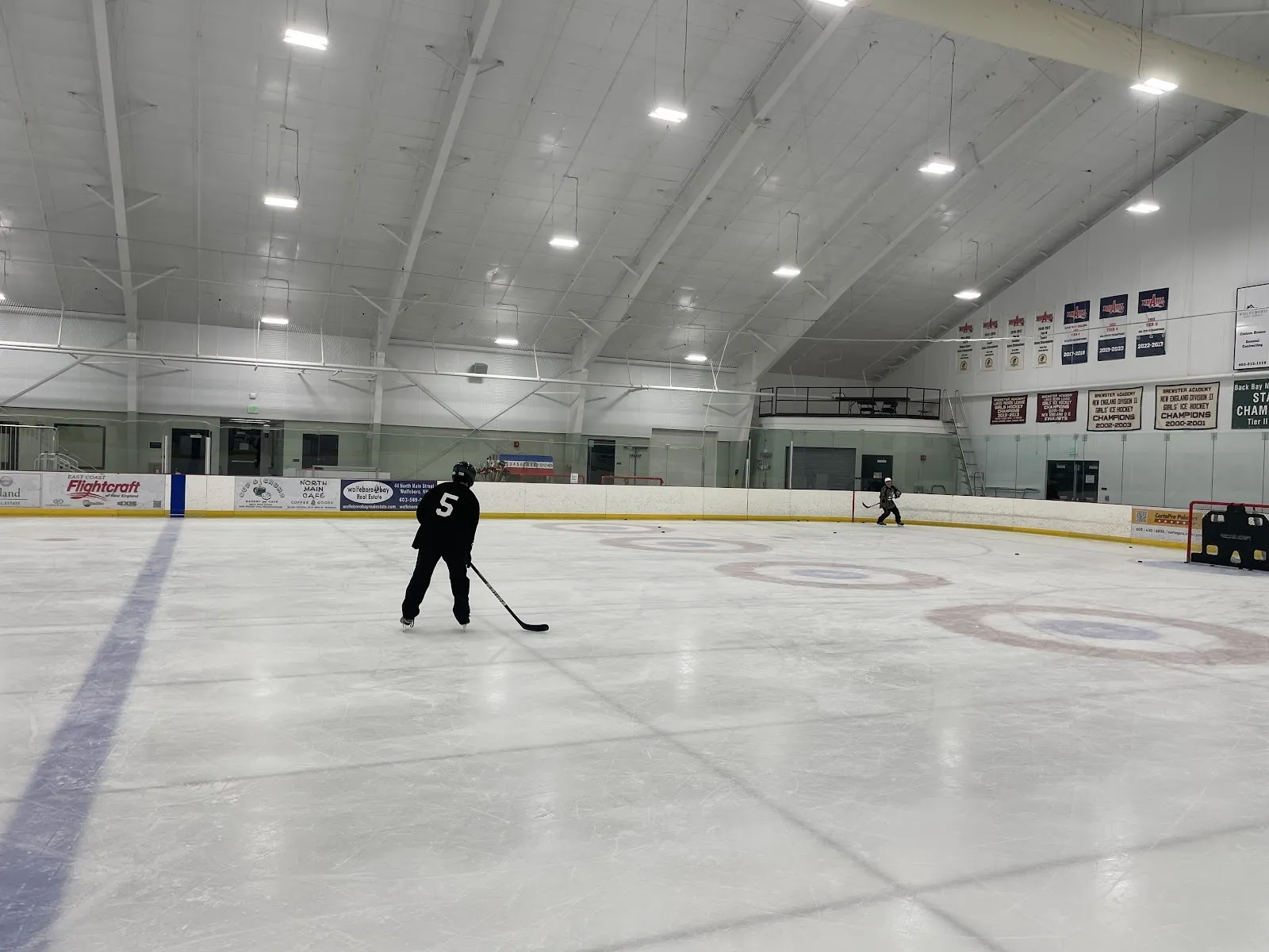 A wide view of the sheet from behind the glass, a player at the left and circles on white ice beneath a bright ceiling with banners on the far wall. — Pop Whalen Ice & Arts Center