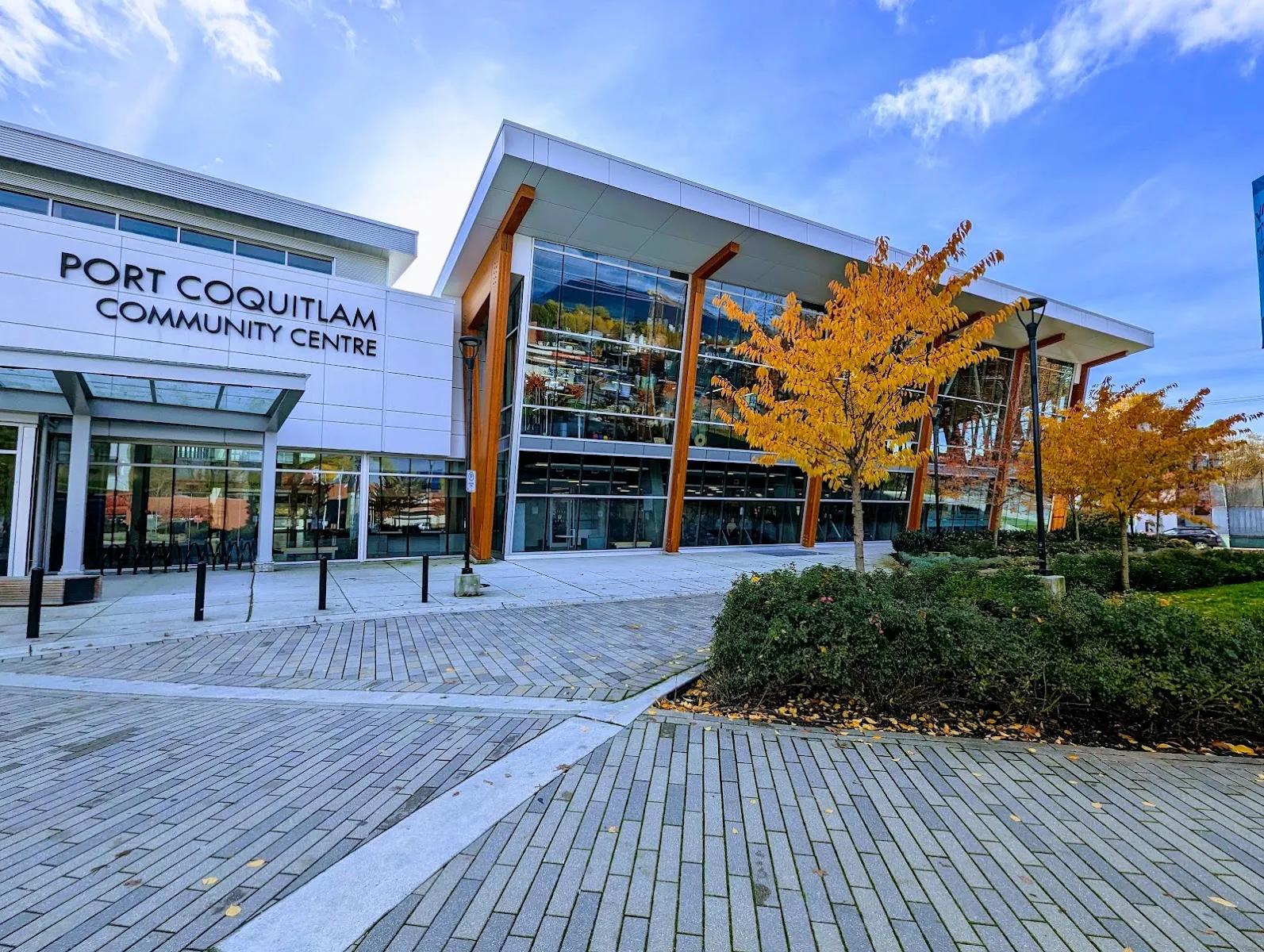 The glass-fronted Port Coquitlam Community Centre with its lettered entrance, autumn trees and a brick plaza. — Port Coquitlam Community Centre