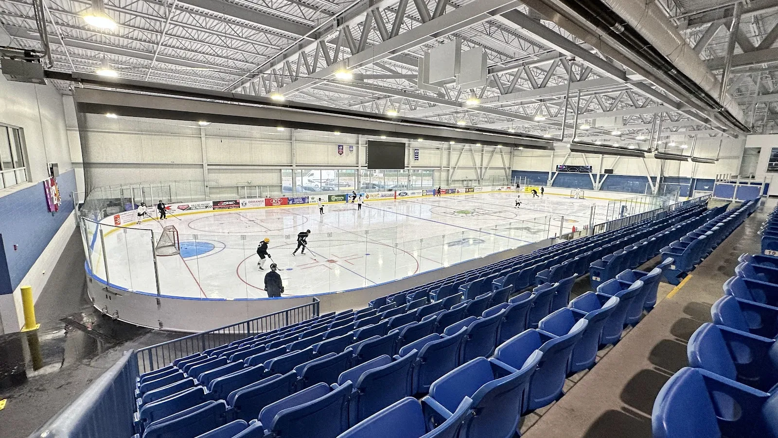 The main arena from the upper seats: rows of blue seats along a sheet under a white steel roof, with a few players on the ice. — Port Coquitlam Community Centre