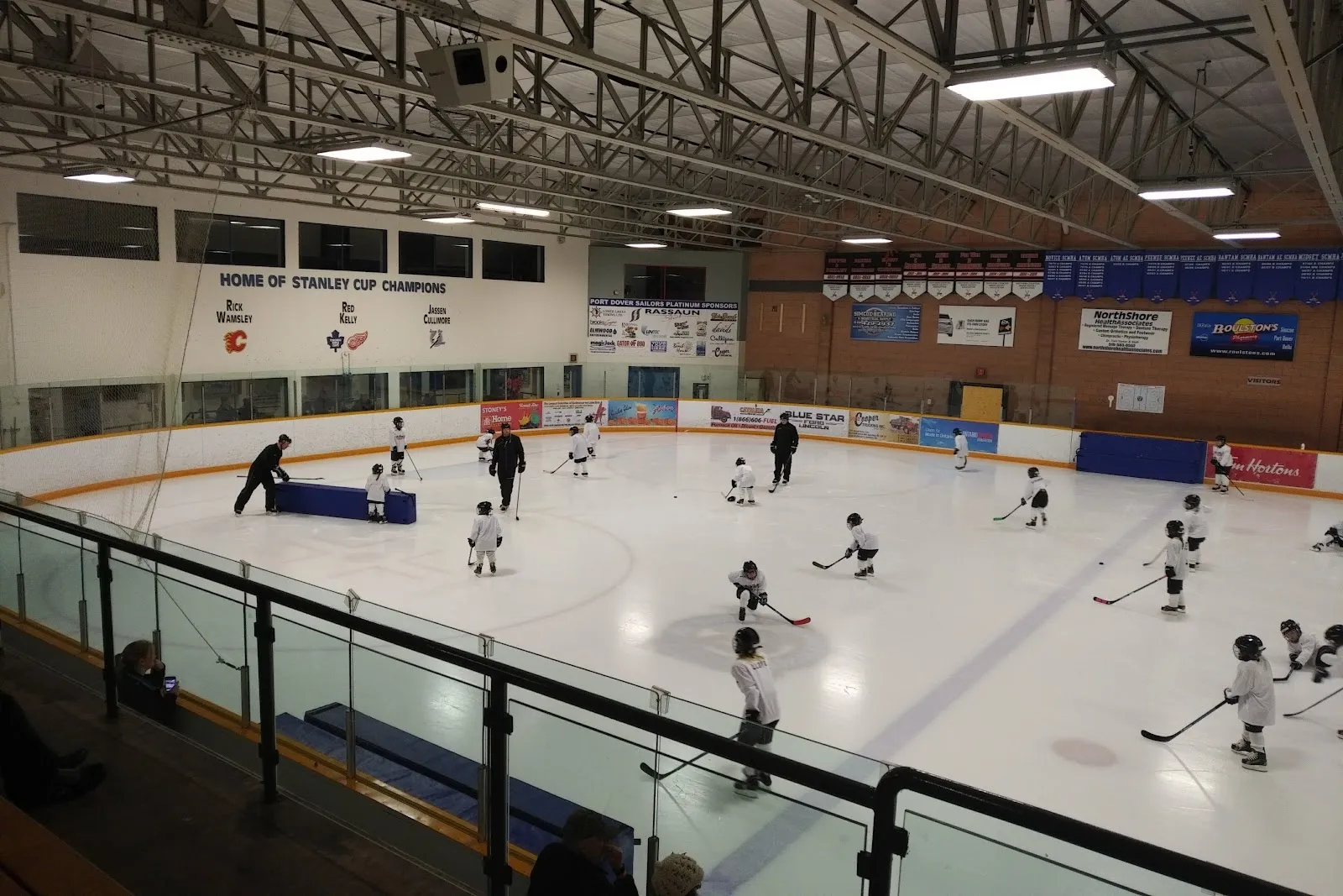 Children skating spread across the ice seen from a glass-fronted viewing rail, steel trusses overhead and club banners along the far wall. — Port Dover Arena