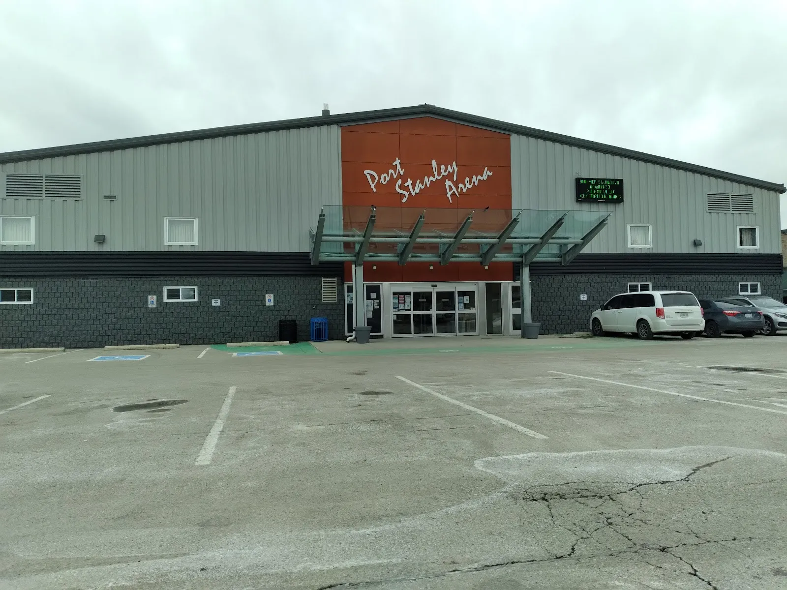 La Port Stanley Arena gris, con su panel con el nombre en letra cursiva naranja y una marquesina de vidrio sobre las puertas de entrada. — Port Stanley Arena & Community Centre