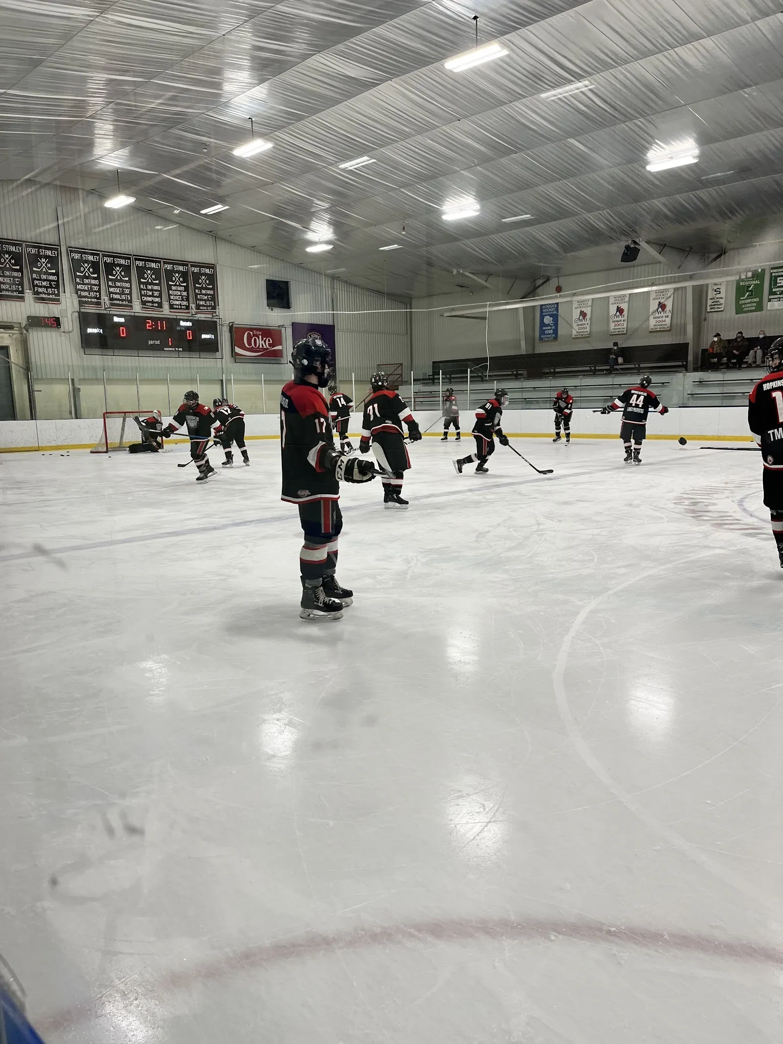 Una vista vertical de la pista con jugadores a lo lejos sobre el hielo y un techo gris con luces sobre las vallas. — Port Stanley Arena & Community Centre