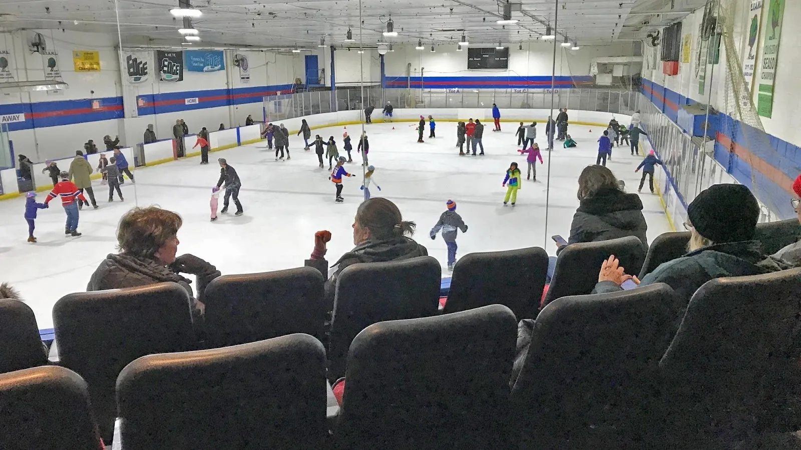 A public session seen from the black seats, skaters spread across a wide sheet with blue-banded boards and a long white wall behind. — Port Washington Skating Center