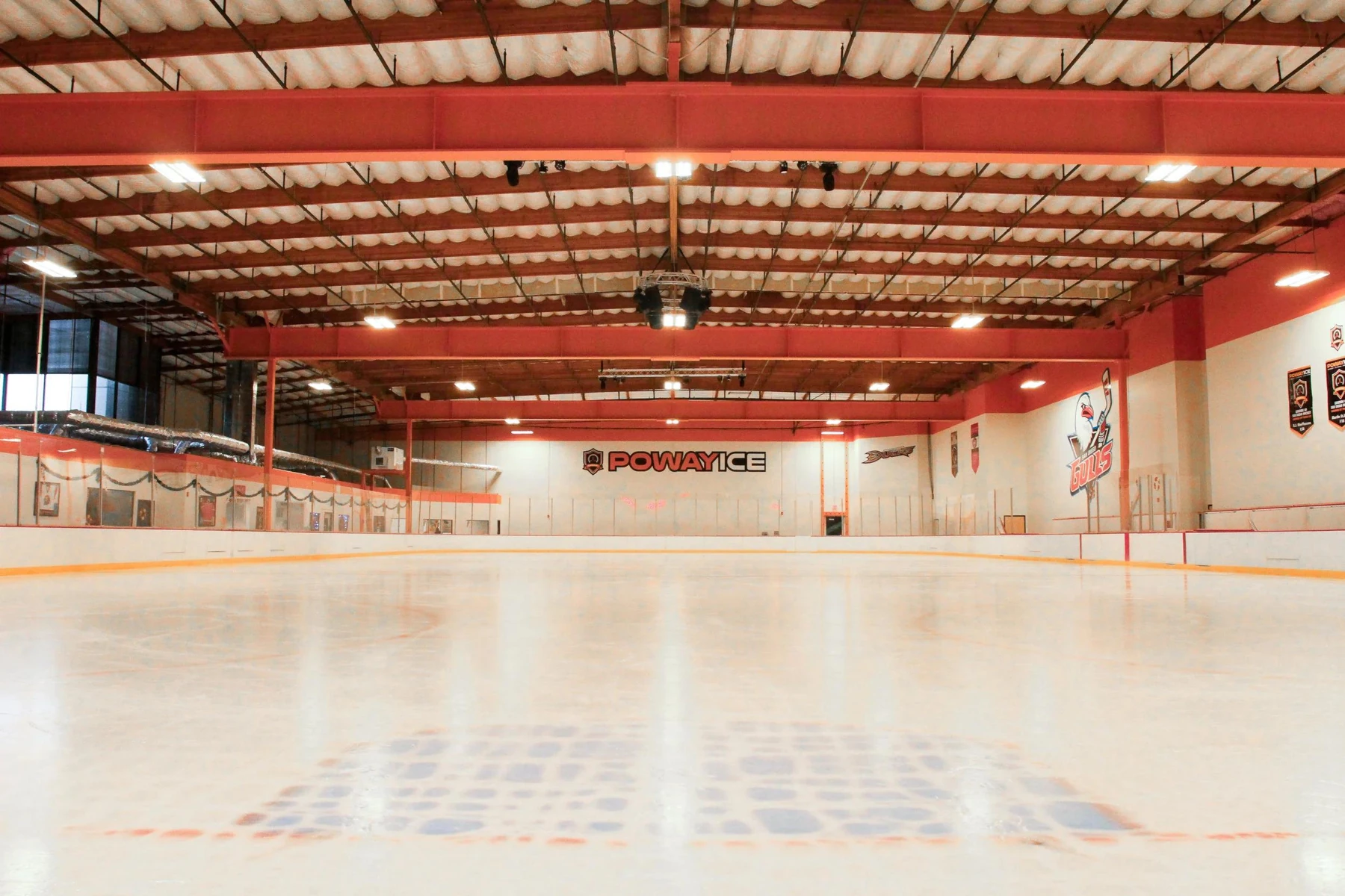 Poway ICE’s empty rink from ice level, beneath red steel roof beams and the Poway Ice sign on the end wall. — Poway ICE