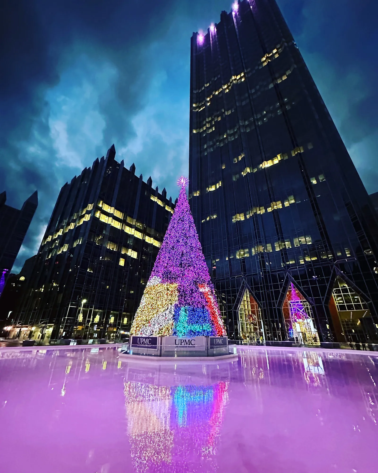 La pista de noche, con su hielo iluminado de morado bajo el árbol de Navidad y las torres de vidrio brillantes de PPG Place. — The UPMC Rink at PPG Place