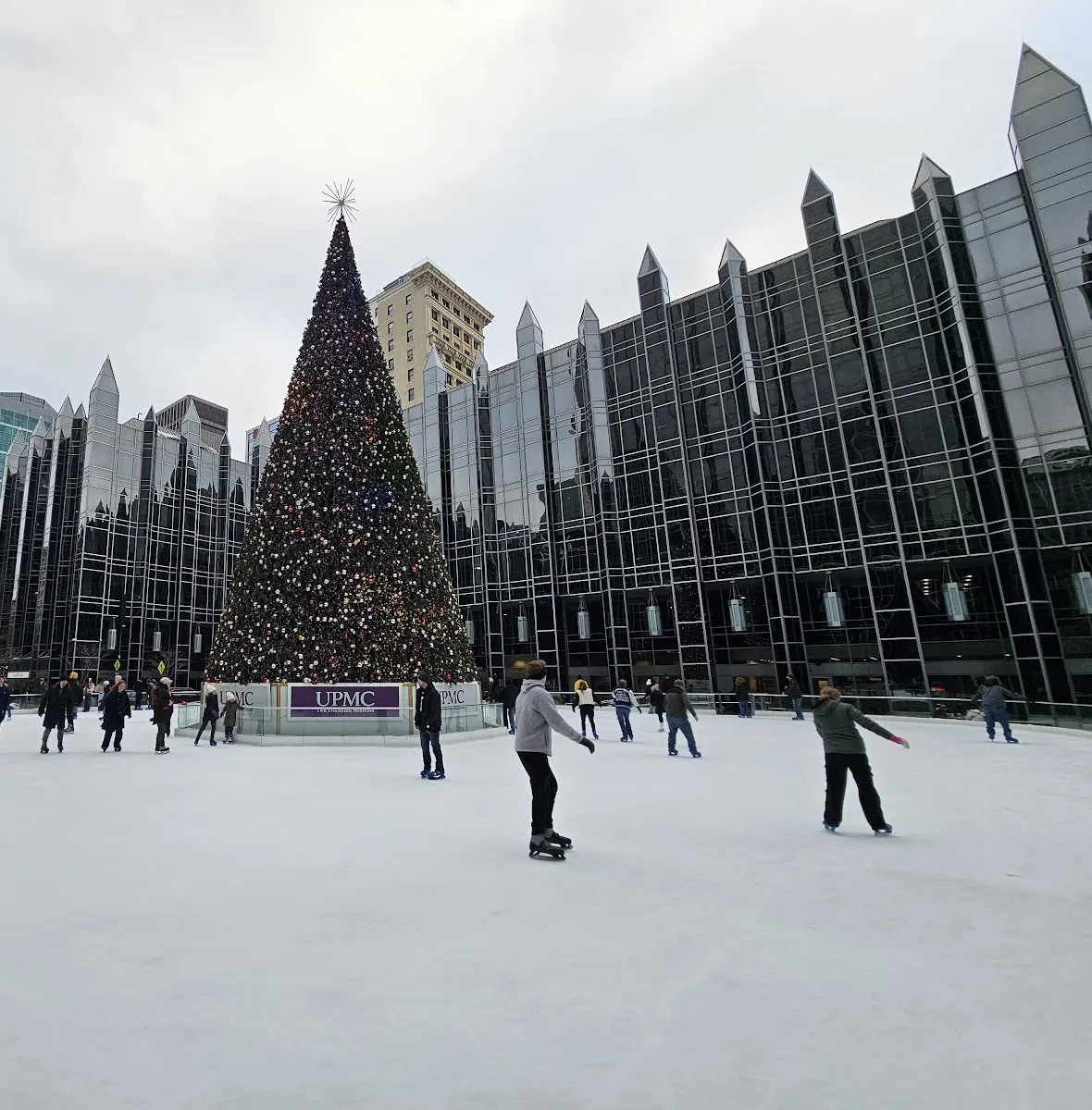 Patinadores dando vueltas en la pista al aire libre bajo el árbol de Navidad iluminado, con las agujas góticas de vidrio de PPG Place detrás. — The UPMC Rink at PPG Place