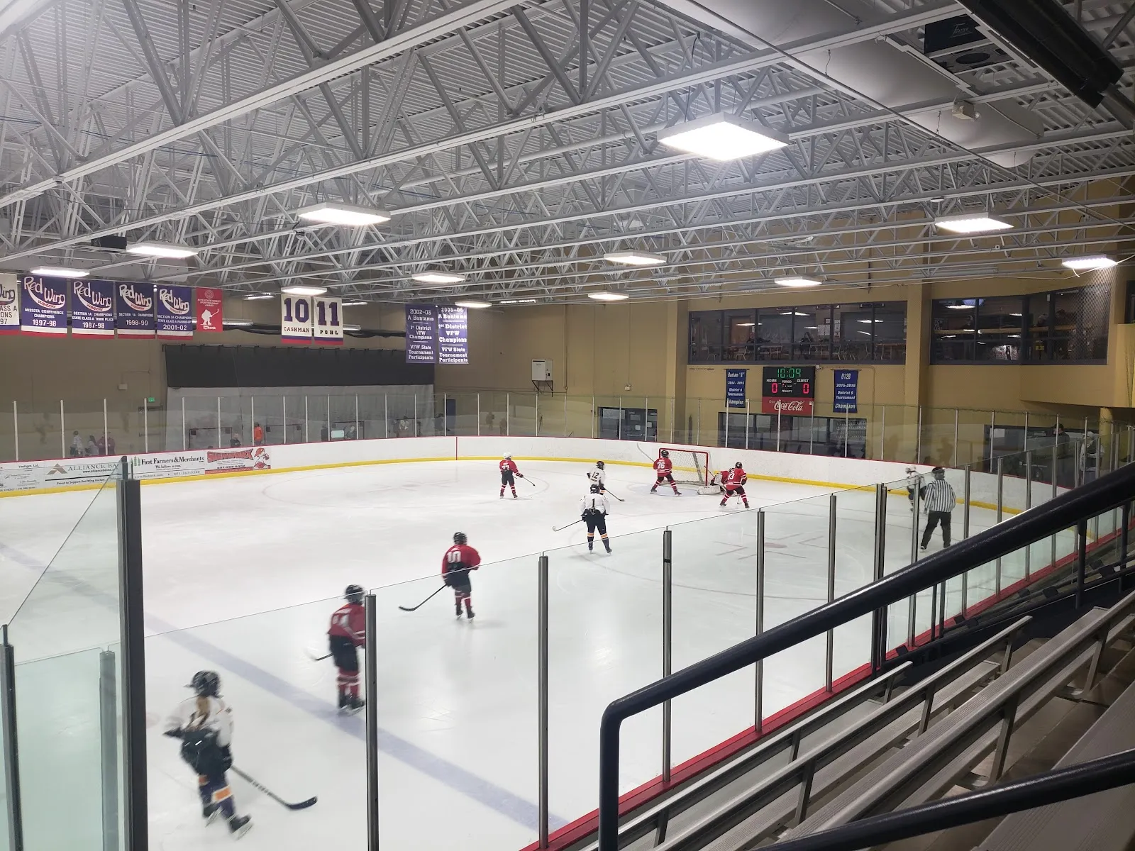The sheet from high in the stands, players spread across the ice under a steel-trussed roof with purple banners hung along the far wall and spectators in the lower rows. — Prairie Island Arena