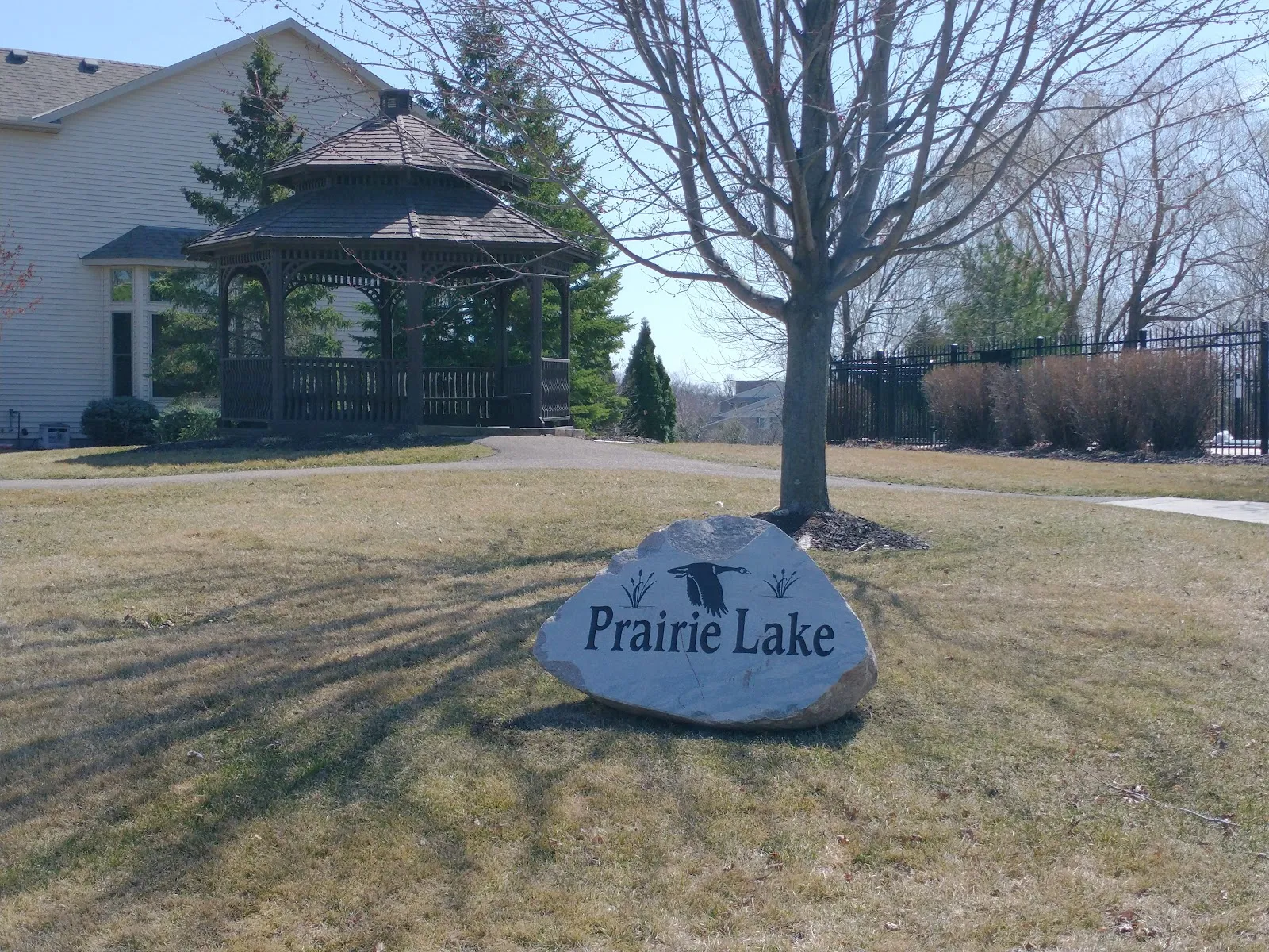 The Prairie Lake park name carved on a boulder beside a path, with a gazebo and a house behind on a bare winter lawn. — Prairie Lake Park Ice Rinks