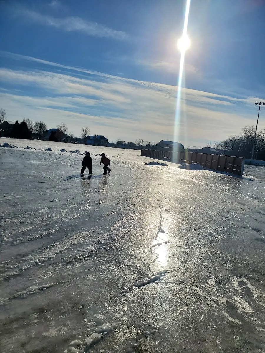 The boarded outdoor rink at Prairie Lake Park on a sunny winter day, with two children on the ice in the distance, the sun low over the ice and light poles and houses beyond. — Prairie Lake Park Ice Rinks