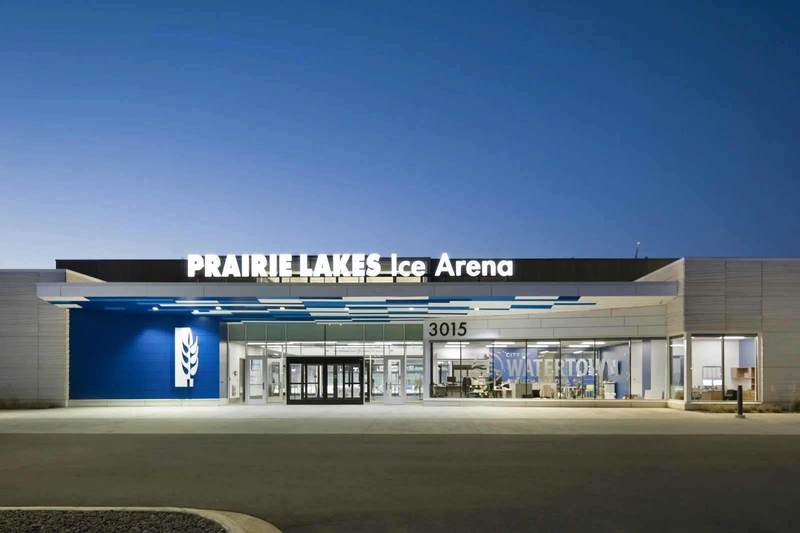 La entrada del Prairie Lakes Ice Arena al atardecer, con su nombre iluminado sobre un vestíbulo de vidrio en el número 3015, junto a un muro azul con un emblema de trigo — Prairie Lakes Ice Arena